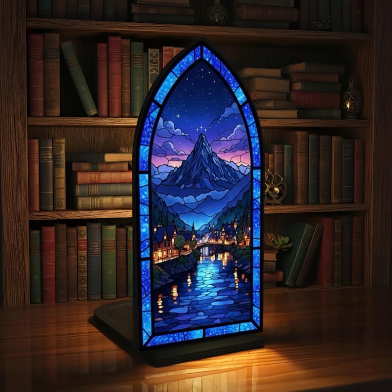 Pu&ograve; includere: Una vetrata raffigurante un paesaggio montano notturno, con un fiume e una citt&agrave; sottostante. La finestra &egrave; incorniciata in blu e poggia su una base nera. Librerie piene di libri sullo sfondo.