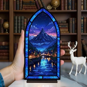Pu&ograve; includere: Un pannello in vetro colorato raffigurante un paesaggio montano con un fiume e una citt&agrave;, incorniciato in blu. L'opera presenta un cielo notturno con stelle e una vetta. Il pannello &egrave; posto su una base di legno.