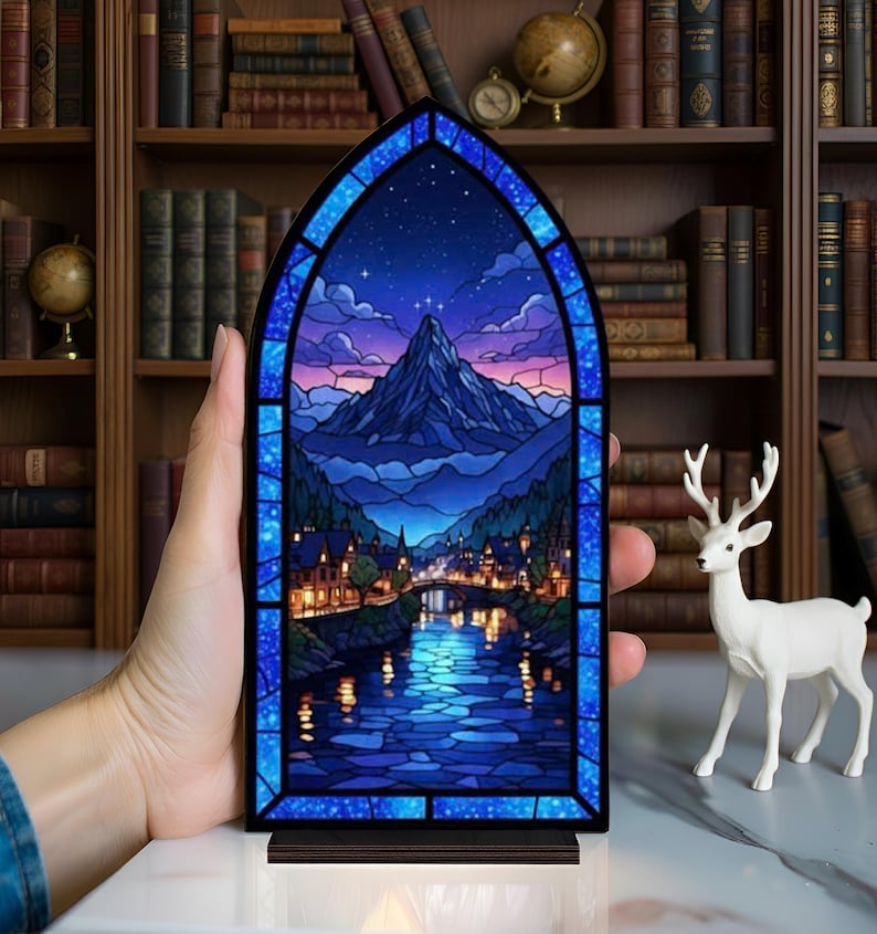 Pu&ograve; includere: Un pannello in vetro colorato raffigurante un paesaggio montano con un fiume e una citt&agrave;, incorniciato in blu. L'opera presenta un cielo notturno con stelle e una vetta. Il pannello &egrave; posto su una base di legno.
