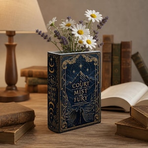 May include: A rectangular dark blue vase shaped like a book titled A Court of Mist and Fury, featuring gold celestial and mountain illustrations. The vase holds a bouquet of white daisies and purple lavender sprigs on a wooden surface.