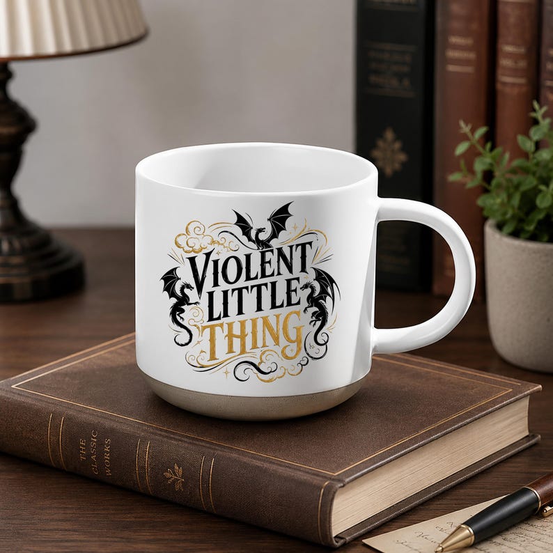 K&ouml;nnte beinhalten: Wei&szlig;e Keramik-Tasse mit dem Text "VIOLENT LITTLE THING" in schwarzer und goldener Schrift, mit Drachen- und Fledermaus-Illustrationen. Die Tasse hat einen grauen Sockel und einen wei&szlig;en Henkel. Sie steht auf einem braunen Buch.