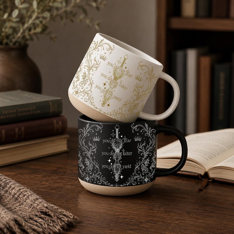 May include: Two ceramic mugs, one white and one black, stacked on a wooden surface. The white mug has gold floral and crown designs with the text "you do not fear, you do not falter, you do not yield." The black mug has similar silver designs and text.