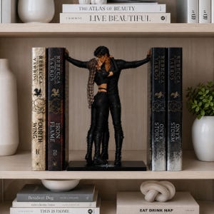 May include: A pair of black bookends featuring a sculpted couple in a close embrace, positioned between two sets of books. The figures are dressed in black attire, with one figure having long braided hair. The bookends rest on a flat black base.