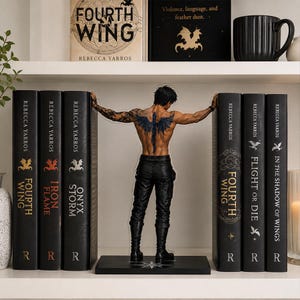 May include: A bookend featuring a male figure with a dark dragon tattoo on his back, wearing black pants and boots, positioned between two stacks of books by Rebecca Yarros, including titles like Fourth Wing, Iron Flame, and Onyx Storm.