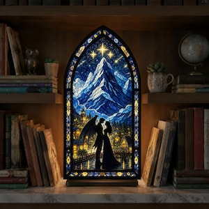 Peut inclure: Une lampe en vitrail en forme d'arche représentant un paysage de montagne bleu sous un ciel étoilé, avec une silhouette ailée et une personne en robe debout sur un pont. Le cadre présente des motifs jaunes et bleus complexes.