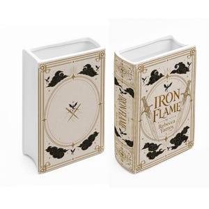 May include: A white ceramic book-shaped vase featuring a cream-colored cover design with gold-toned sword illustrations, black cloud motifs, and small dragon silhouettes. The front displays the text Iron Flame by Rebecca Yarros in gold lettering.