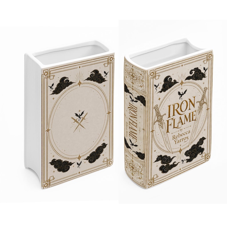 May include: A white ceramic book-shaped vase featuring a cream-colored cover design with gold-toned sword illustrations, black cloud motifs, and small dragon silhouettes. The front displays the text Iron Flame by Rebecca Yarros in gold lettering.
