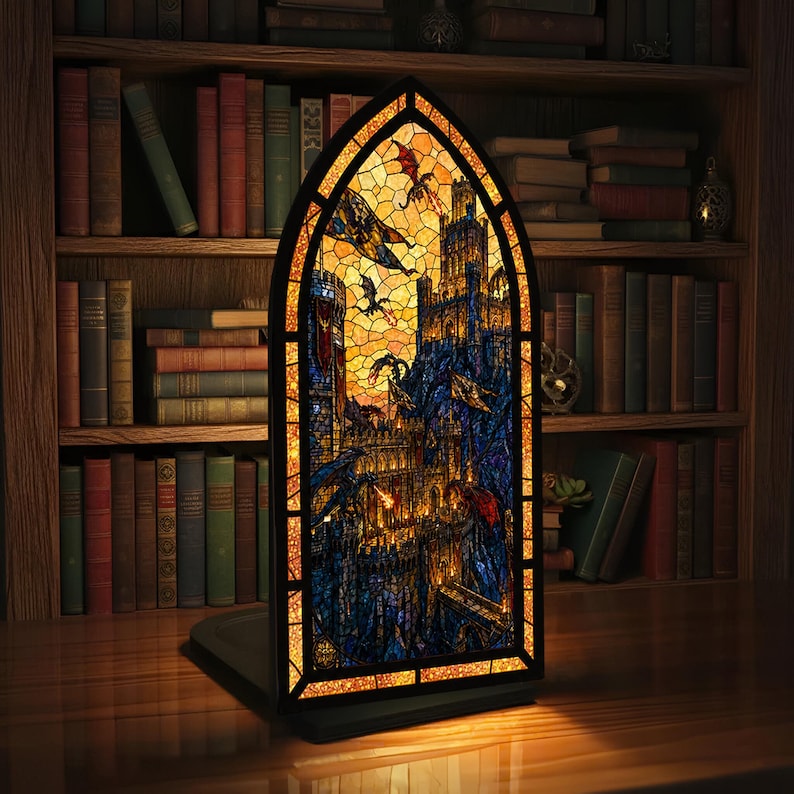 May include: A stained glass window showing a fantasy castle scene with dragons, in shades of blue, orange, and yellow. The window is arched and framed in gold, with a black base. The window is illuminated, creating a warm glow.