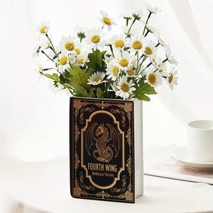 Puede incluir: Un jarr&oacute;n blanco con forma de libro con el t&iacute;tulo "FOURTH WING" y una ilustraci&oacute;n de drag&oacute;n dorada. Contiene un ramo de margaritas blancas con centros amarillos y hojas verdes.