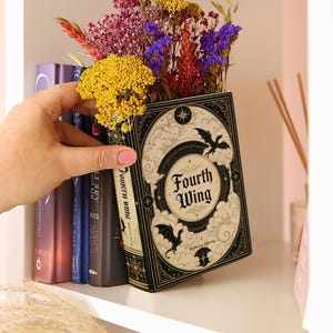 May include: A decorative book-shaped planter titled Fourth Wing by Rebecca Yarros, featuring black and cream ornate illustrations of dragons, clouds, and stars. The hollow interior holds a vibrant arrangement of dried yellow, purple, and red flowers.