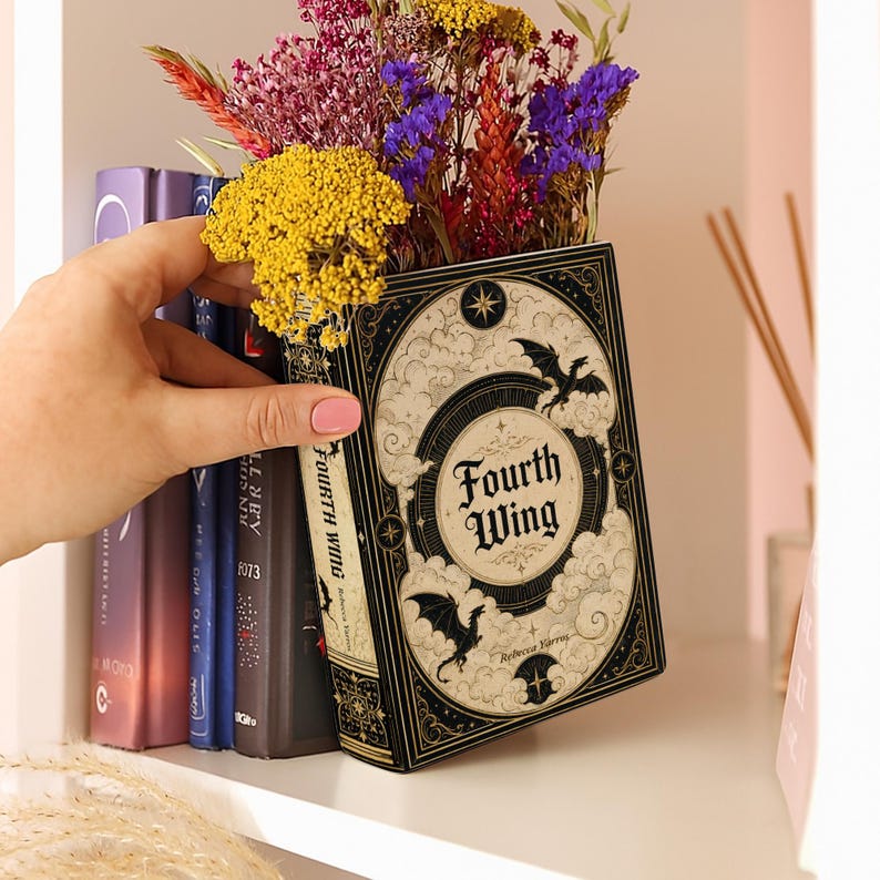 May include: A decorative book-shaped planter titled Fourth Wing by Rebecca Yarros, featuring black and cream ornate illustrations of dragons, clouds, and stars. The hollow interior holds a vibrant arrangement of dried yellow, purple, and red flowers.