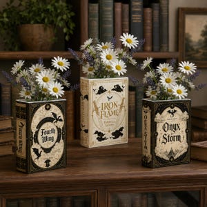 Pu&ograve; includere: Tre vasi rettangolari a forma di libro con copertine nere e crema con i titoli Fourth Wing, Iron Flame e Onyx Storm. Ogni vaso &egrave; riempito con fiori bianchi simili a margherite e rametti viola, disposti su una superficie in legno.
