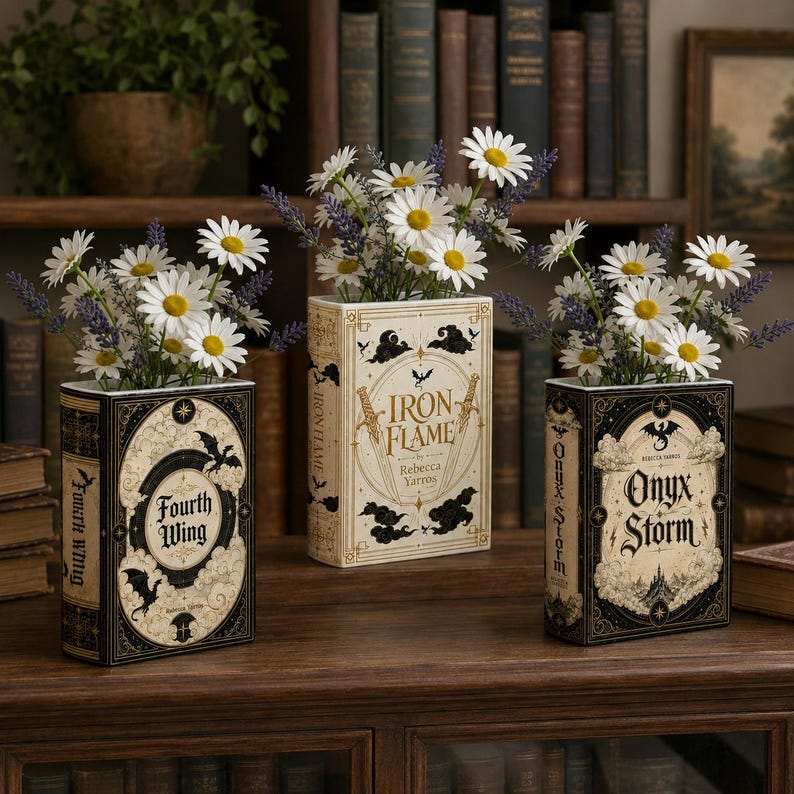 Pu&ograve; includere: Tre vasi rettangolari a forma di libro con copertine nere e crema con i titoli Fourth Wing, Iron Flame e Onyx Storm. Ogni vaso &egrave; riempito con fiori bianchi simili a margherite e rametti viola, disposti su una superficie in legno.