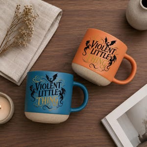 K&ouml;nnte beinhalten: Zwei Keramikbecher, einer orange und einer blau, mit dem Text "VIOLENT LITTLE THING" in schwarzer und goldener Schrift. Jeder Becher hat einen beigen Boden und einen passenden Henkel. Die Becher stehen auf einer Holzoberfl&auml;che.