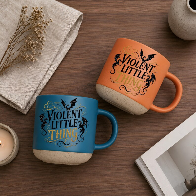 K&ouml;nnte beinhalten: Zwei Keramikbecher, einer orange und einer blau, mit dem Text "VIOLENT LITTLE THING" in schwarzer und goldener Schrift. Jeder Becher hat einen beigen Boden und einen passenden Henkel. Die Becher stehen auf einer Holzoberfl&auml;che.