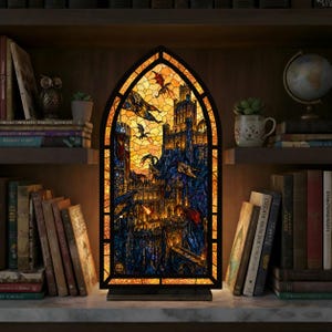 Könnte beinhalten: Eine Buntglaslampe mit einer Fantasy-Burgszene mit Drachen in Orange-, Blau- und Gelbtönen. Die Lampe hat einen schwarzen Rahmen und einen Holzsockel. Sie steht auf einer Marmoroberfläche, umgeben von Büchern.