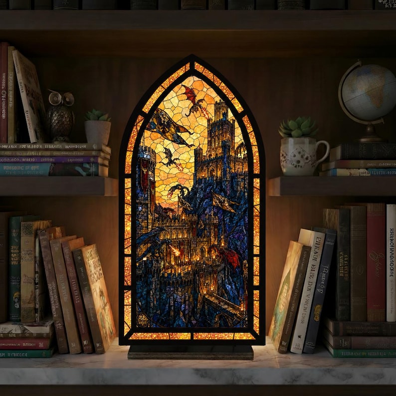 May include: A gothic-style stained glass lamp featuring a fantasy castle scene with dragons, in orange, blue, and yellow hues. The lamp has a black frame and a wooden base. It is placed on a marble surface, surrounded by books on a bookshelf.