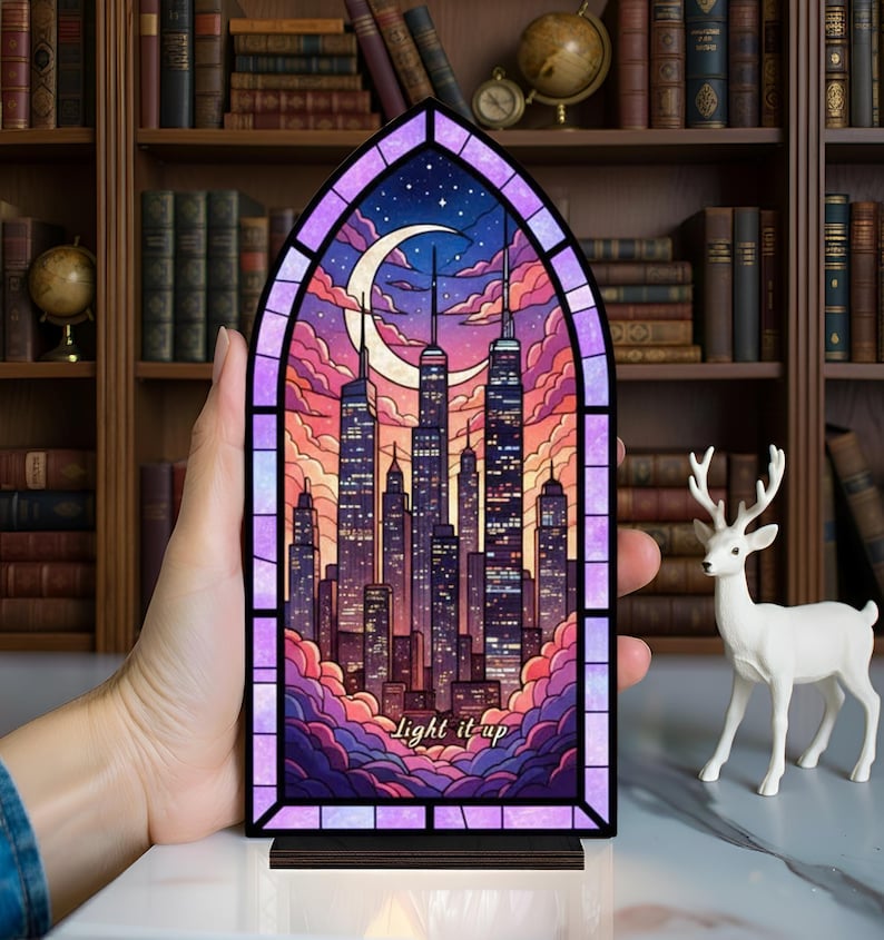 May include: A stained glass panel depicting a cityscape at night, with a crescent moon and stars. The panel is framed in purple and has the words "Light it up" at the bottom. The panel is set on a wooden base.