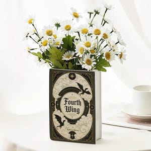May include: A rectangular ceramic vase shaped like a book, featuring a cream-colored cover with black and gold illustrations of dragons, clouds, and stars. The text "Fourth Wing" is printed on the front. It is filled with white daisy flowers.
