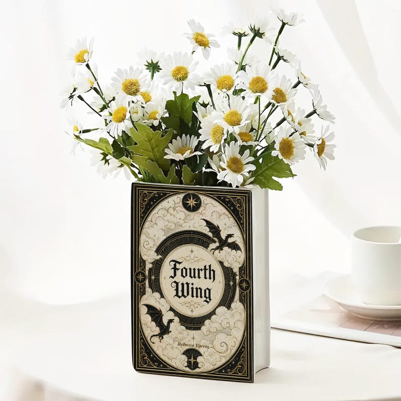 May include: A rectangular ceramic vase shaped like a book, featuring a cream-colored cover with black and gold illustrations of dragons, clouds, and stars. The text "Fourth Wing" is printed on the front. It is filled with white daisy flowers.