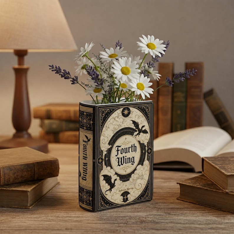 Pu&ograve; includere: Un vaso rettangolare a forma di libro con un design di copertina color crema e nero con il testo Fourth Wing, decorato con fiori di margherita bianchi e rametti di lavanda viola. Il vaso &egrave; posizionato su una superficie di legno tra altri libri.