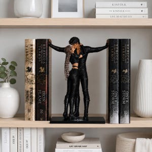 May include: A pair of black bookends featuring a sculpted couple in a close embrace, positioned between two sets of books. The figures are dressed in black outfits, with one figure having long braided hair. The book spines display the text Rebecca Yarros.