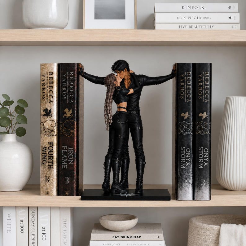 May include: A pair of black bookends featuring a sculpted couple in a close embrace, positioned between two sets of books. The figures are dressed in black outfits, with one figure having long braided hair. The book spines display the text Rebecca Yarros.