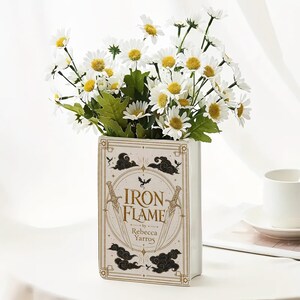 May include: A rectangular ceramic vase shaped like a book with the title Iron Flame by Rebecca Yarros printed on the front. The design features gold swords, black clouds, and a small dragon silhouette. It is filled with white daisies with yellow centers.