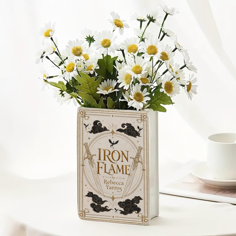 May include: A rectangular ceramic vase shaped like a book with the title Iron Flame by Rebecca Yarros printed on the front. The design features gold swords, black clouds, and a small dragon silhouette. It is filled with white daisies with yellow centers.