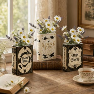 Pu&ograve; includere: Tre vasi rettangolari a forma di libro con i titoli Fourth Wing, Iron Flame e Onyx Storm. Ogni vaso &egrave; riempito con fiori bianchi simili a margherite e rametti di lavanda viola, disposti su una superficie in legno vicino a una finestra.