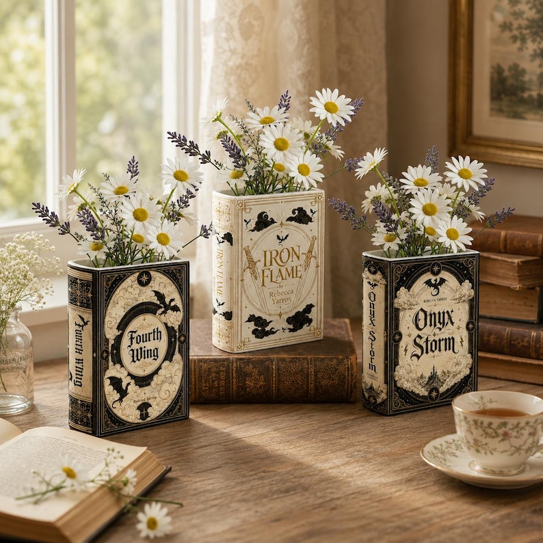 Pu&ograve; includere: Tre vasi rettangolari a forma di libro con i titoli Fourth Wing, Iron Flame e Onyx Storm. Ogni vaso &egrave; riempito con fiori bianchi simili a margherite e rametti di lavanda viola, disposti su una superficie in legno vicino a una finestra.