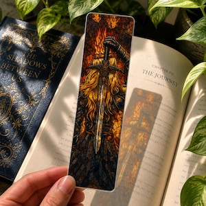 May include: A rectangular bookmark featuring a stained-glass style illustration of a figure with long golden hair holding a large sword upright. The background displays orange and yellow flames, while the figure wears dark armour with intricate details.