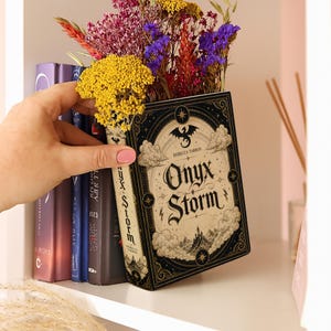 Puede incluir: Un libro titulado Onyx Storm de Rebecca Yarros con una portada negra y crema que muestra un drag&oacute;n, nubes y un castillo, que funciona como maceta llena de un ramo de flores secas amarillas, moradas, rojas y naranjas.
