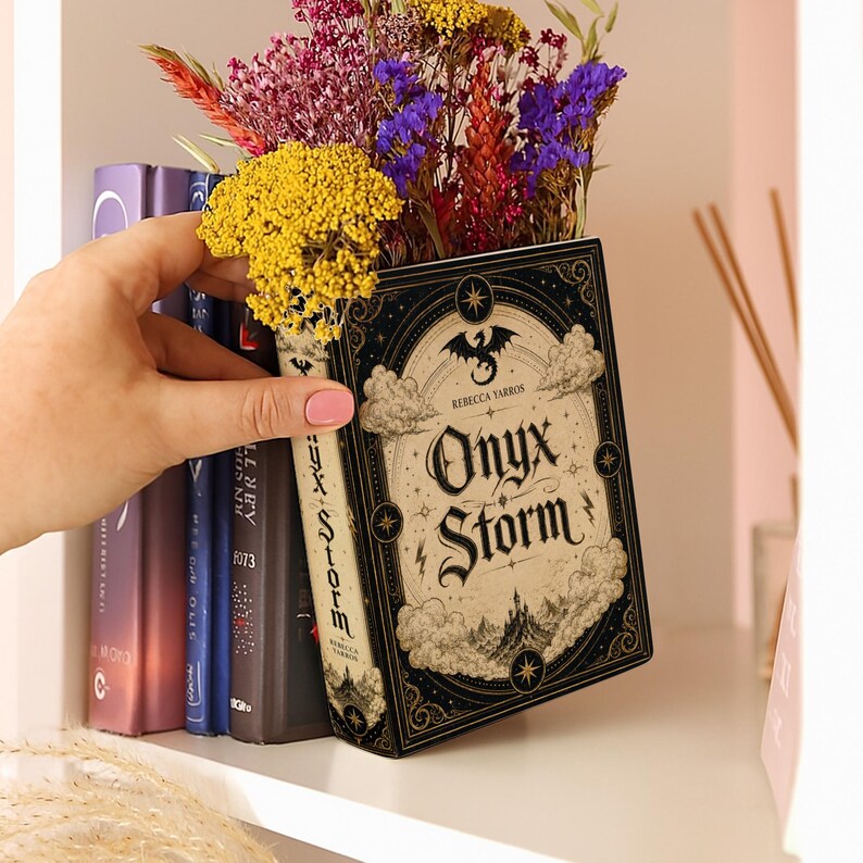 Puede incluir: Un libro titulado Onyx Storm de Rebecca Yarros con una portada negra y crema que muestra un drag&oacute;n, nubes y un castillo, que funciona como maceta llena de un ramo de flores secas amarillas, moradas, rojas y naranjas.