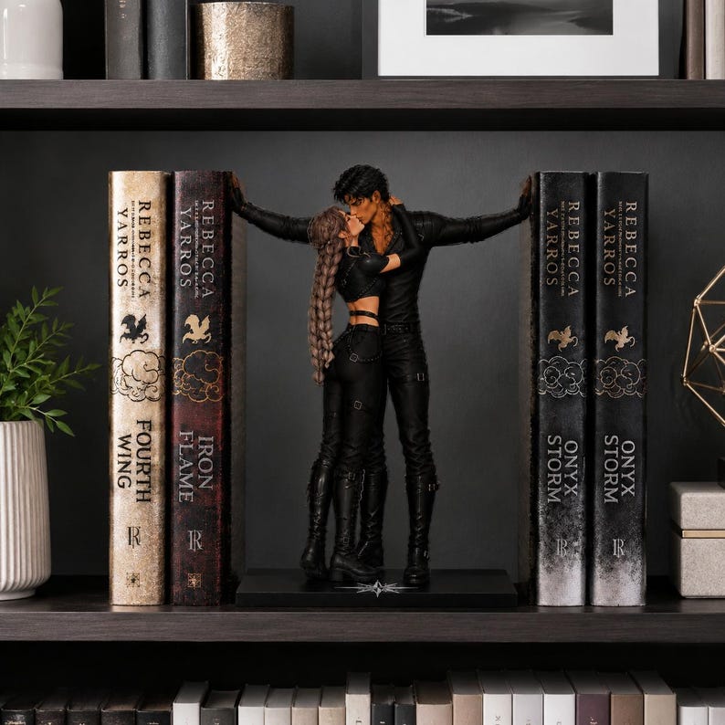 May include: A decorative bookend set featuring a sculpted pair of figures in black attire embracing between two stacks of books. The books include titles by Rebecca Yarros such as Fourth Wing, Iron Flame, and Onyx Storm, with gold lettering on spines.