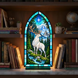 Op de afbeelding: Een boogvormige glas-in-loodlamp met een wit hert in een bos met een berg, een kasteel en een heldere ster aan de hemel. De lamp heeft een zwart frame, staat op een voet en verspreidt een zachte gloed vanuit het glazen paneel.