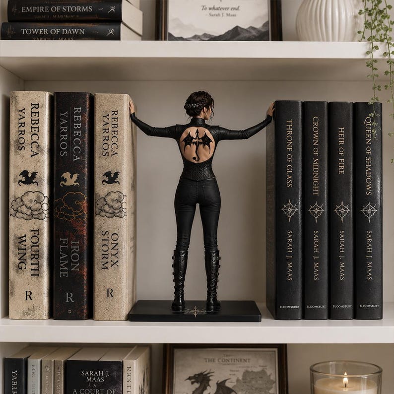 May include: A figurine of a person with dark hair in a braided updo, wearing a black long-sleeved top with a dragon tattoo on the back and black pants, standing between two rows of books on a shelf, with hands touching the book spines.