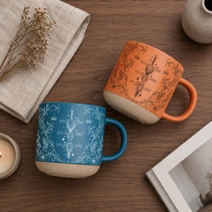 May include: Two ceramic mugs with handles. One is orange, the other is teal. Both mugs have a beige base and feature a sword design with the text "you do not fear, you do not falter, you do not yield."