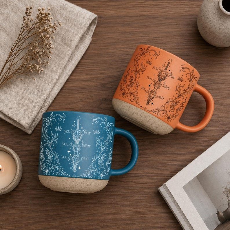 May include: Two ceramic mugs with handles. One is orange, the other is teal. Both mugs have a beige base and feature a sword design with the text "you do not fear, you do not falter, you do not yield."