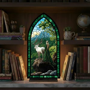 May include: A decorative stained glass lamp depicting a white deer with antlers in a mountain scene. The lamp has a green border and is lit, creating a warm glow. The lamp is displayed on a shelf with books and is a decorative item.
