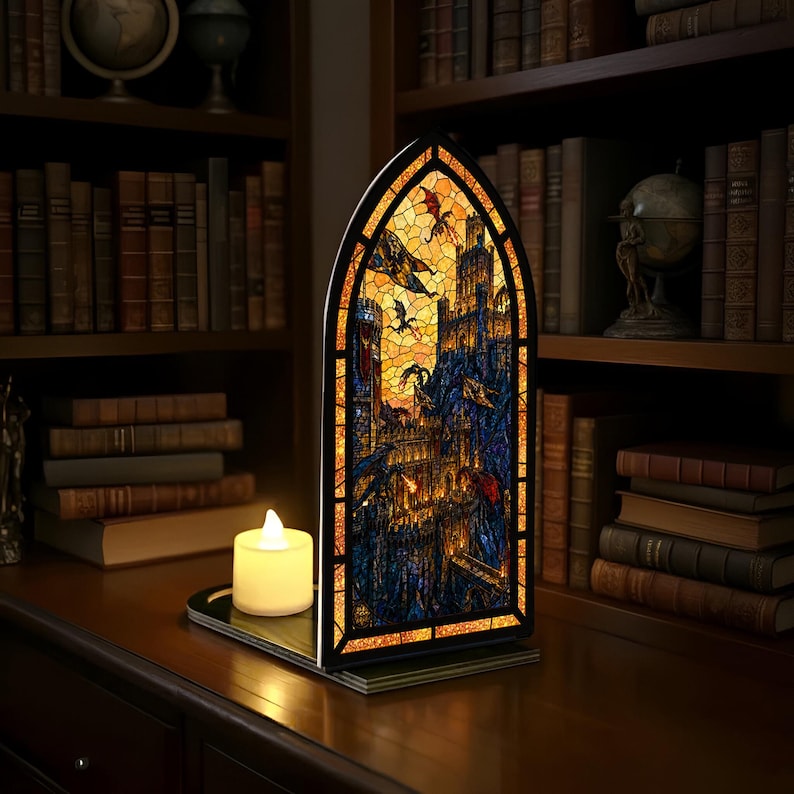 May include: A decorative stained glass panel showing a fantasy castle scene with dragons, lit from behind. The panel has an arched top and sits on a black base. A battery-powered candle is in front of the panel.