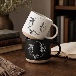 May include: Two ceramic mugs stacked vertically, one white with a black dragon design and one black with a white dragon design. Both mugs feature a circular pattern of three flying dragons surrounded by small stars and dots, with a natural tan base.