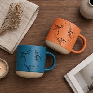 Op de afbeelding: Twee keramische mokken, &eacute;&eacute;n teal en &eacute;&eacute;n oranje, elk met een drakenontwerp. De mokken hebben een matte afwerking en een lichtgekleurde basis. De drakenillustratie is zwart en omcirkelt de mok.