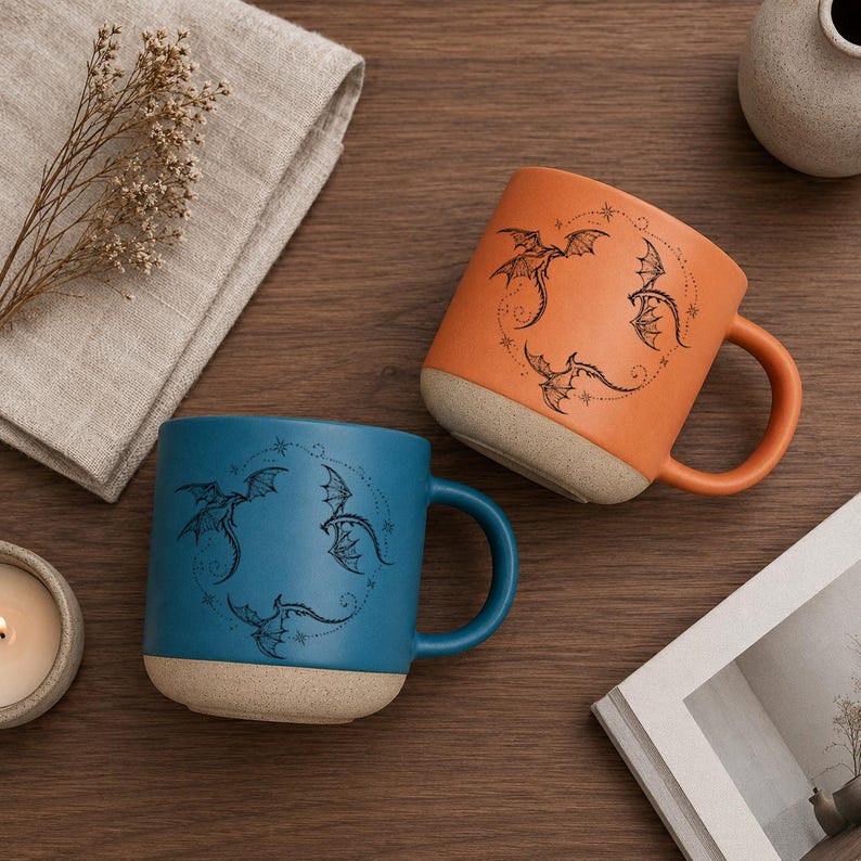 Op de afbeelding: Twee keramische mokken, &eacute;&eacute;n teal en &eacute;&eacute;n oranje, elk met een drakenontwerp. De mokken hebben een matte afwerking en een lichtgekleurde basis. De drakenillustratie is zwart en omcirkelt de mok.