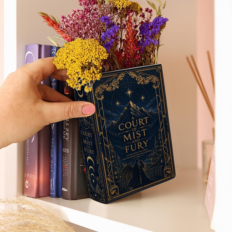 May include: A rectangular book-shaped vase in dark blue with gold filigree, featuring the title A Court of Mist and Fury, a mountain landscape, and a couple silhouette. The vase is filled with a bouquet of dried yellow, purple, and red flowers.