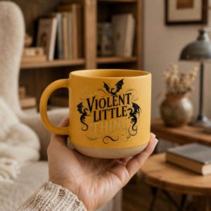 K&ouml;nnte beinhalten: Senfgelbe Keramik-Tasse mit dem Schriftzug "Violent Little Thing" in schwarzer Schrift und Drachenillustrationen. Die Tasse hat einen beigefarbenen Boden und einen gebogenen Henkel. Die Tasse wird in einer Hand gehalten.