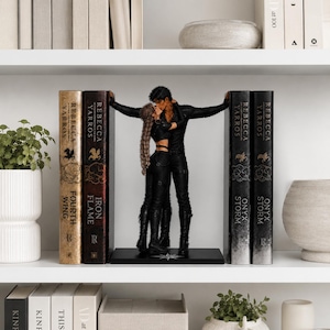 May include: A black bookend set featuring two figures in black clothing embracing and kissing, positioned between two pairs of books titled Fourth Wing, Iron Flame, Onyx Storm, and Onyx Storm by Rebecca Yarros on a white shelf.