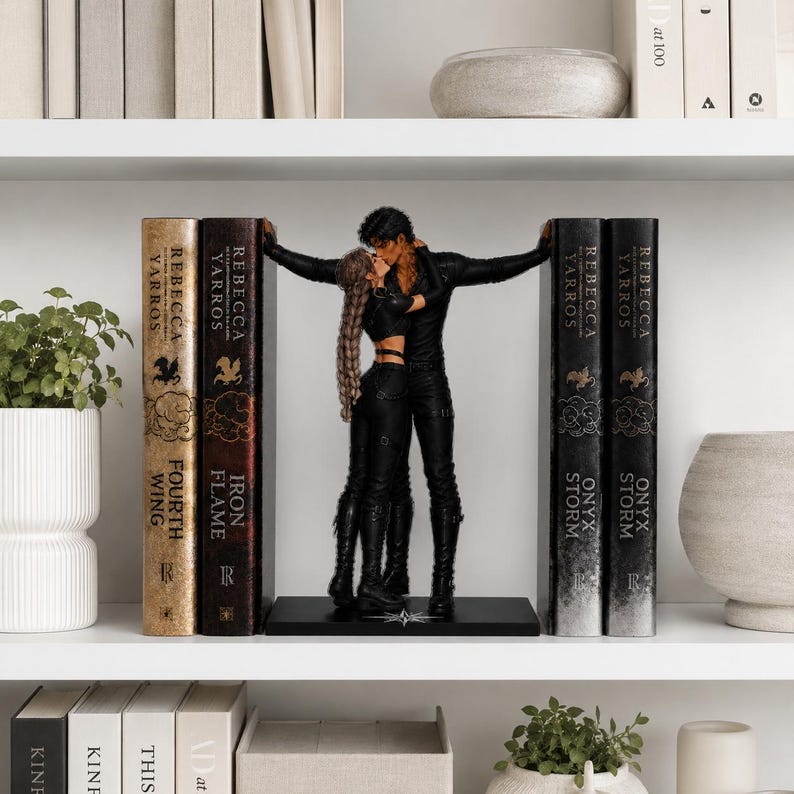 May include: A black bookend set featuring two figures in black clothing embracing and kissing, positioned between two pairs of books titled Fourth Wing, Iron Flame, Onyx Storm, and Onyx Storm by Rebecca Yarros on a white shelf.
