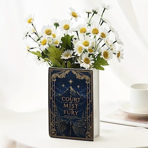 May include: A rectangular ceramic vase shaped like a book with a dark blue cover featuring gold filigree, a mountain illustration, and the text A Court of Mist and Fury. The vase is filled with a bouquet of white daisies with yellow centers.