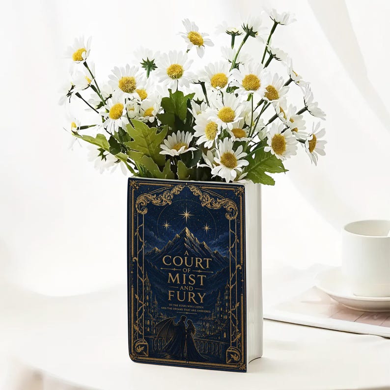 May include: A rectangular ceramic vase shaped like a book with a dark blue cover featuring gold filigree, a mountain illustration, and the text A Court of Mist and Fury. The vase is filled with a bouquet of white daisies with yellow centers.